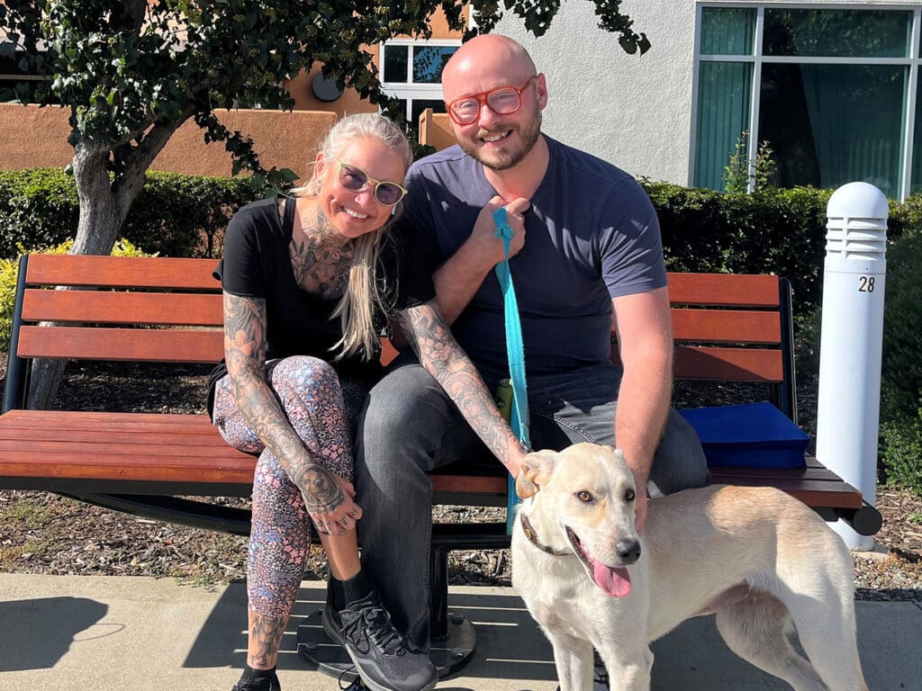 A man and a woman sitting on a park bench, smiling with a dog beside them. The woman has long, light-colored hair and tattoos, wearing sunglasses and patterned leggings. The man, with a shaved head and glasses, is holding the dog's leash. The setting appears sunny, with landscaping in the background. This image relates to Adoptions at Joybound.