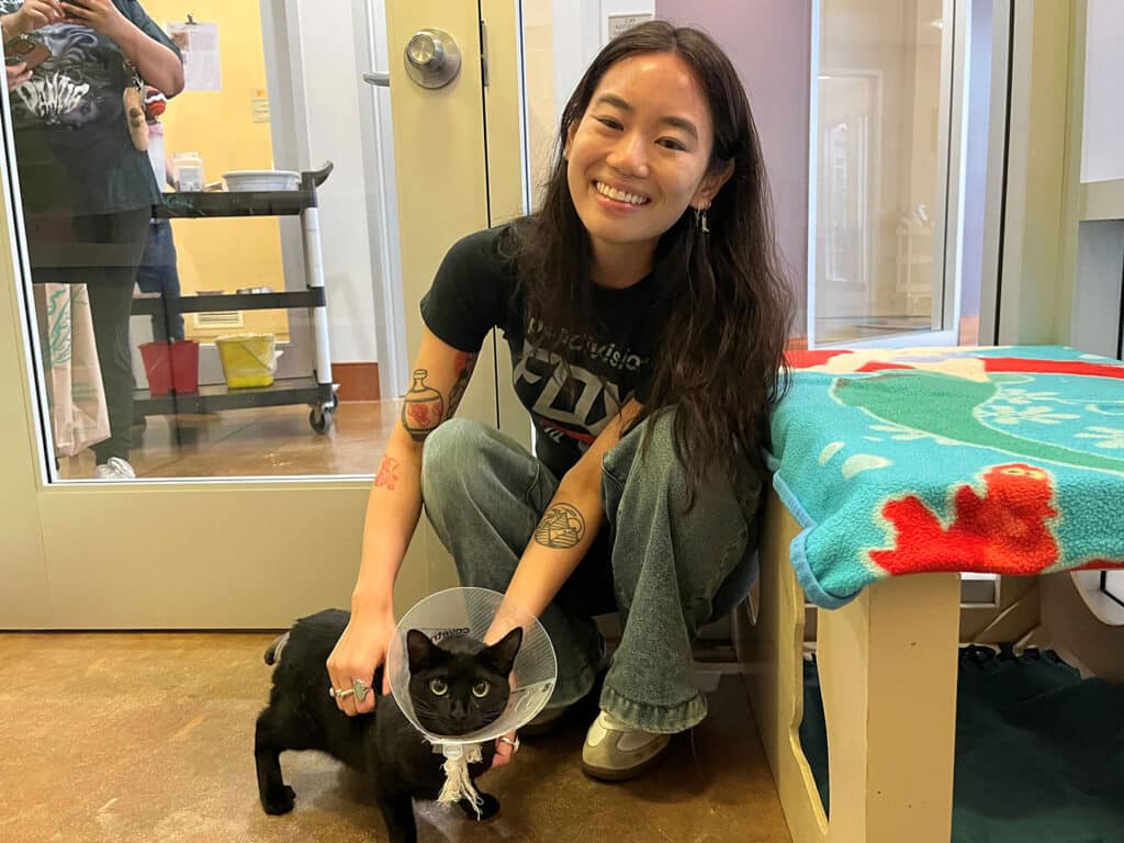 A smiling woman with long hair sits beside a black cat wearing an Elizabethan collar in an animal shelter. The background features a grooming area and colorful blankets. This scene is related to Adoptions at Joybound.