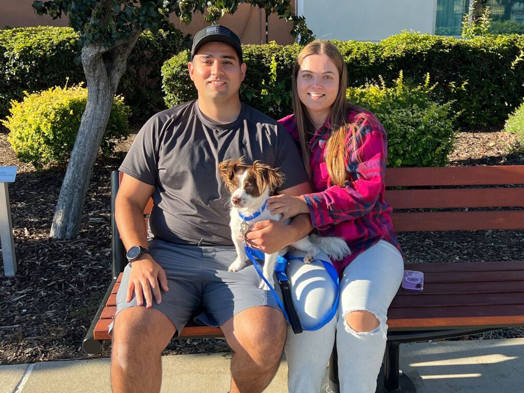 A man and woman sitting on a bench in a park, both smiling as they hold a small dog. The man is wearing a dark t-shirt and shorts, while the woman is in a plaid shirt. Surrounding greenery is visible, contributing to a pleasant setting. Adoptions at Joybound.