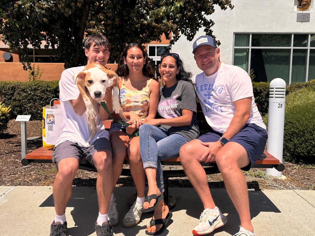 A family of four sits on a bench outside, smiling. A young man holds a dog on his lap, while two women and another man pose beside him. In the background, greenery and a building are visible. This image relates to "Adoptions at Joybound."