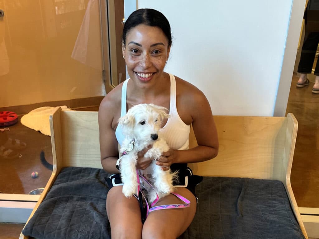 A woman with dark hair in a white tank top sits on a wooden bench, holding a small white dog on a leash. The background shows a pet adoption setting with toys and other pets visible. The image relates to "Adoptions at Joybound".