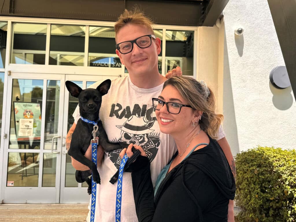 A man and a woman stand outside a building, smiling while holding a small black dog on a leash. The man wears glasses and a graphic t-shirt, and the woman has glasses and a black jacket. The image represents "Adoptions at Joybound".