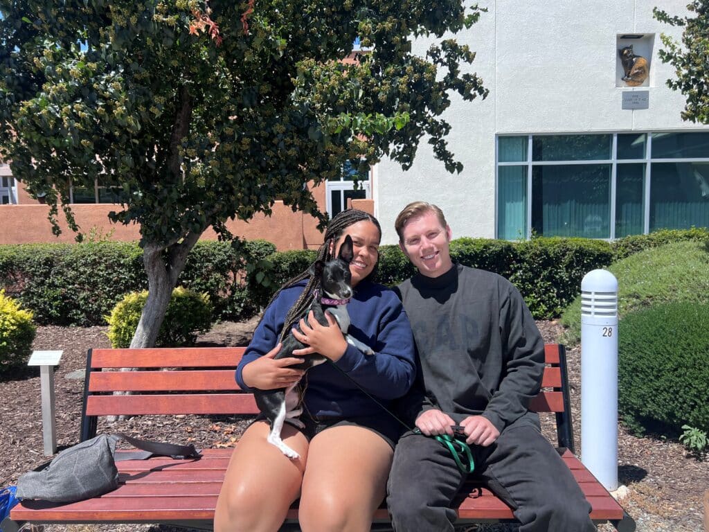 A woman and a man sitting on a wooden bench outdoors, smiling while holding a small dog. The background includes greenery and a building, suggesting a public space. The sun is shining, creating a bright atmosphere.