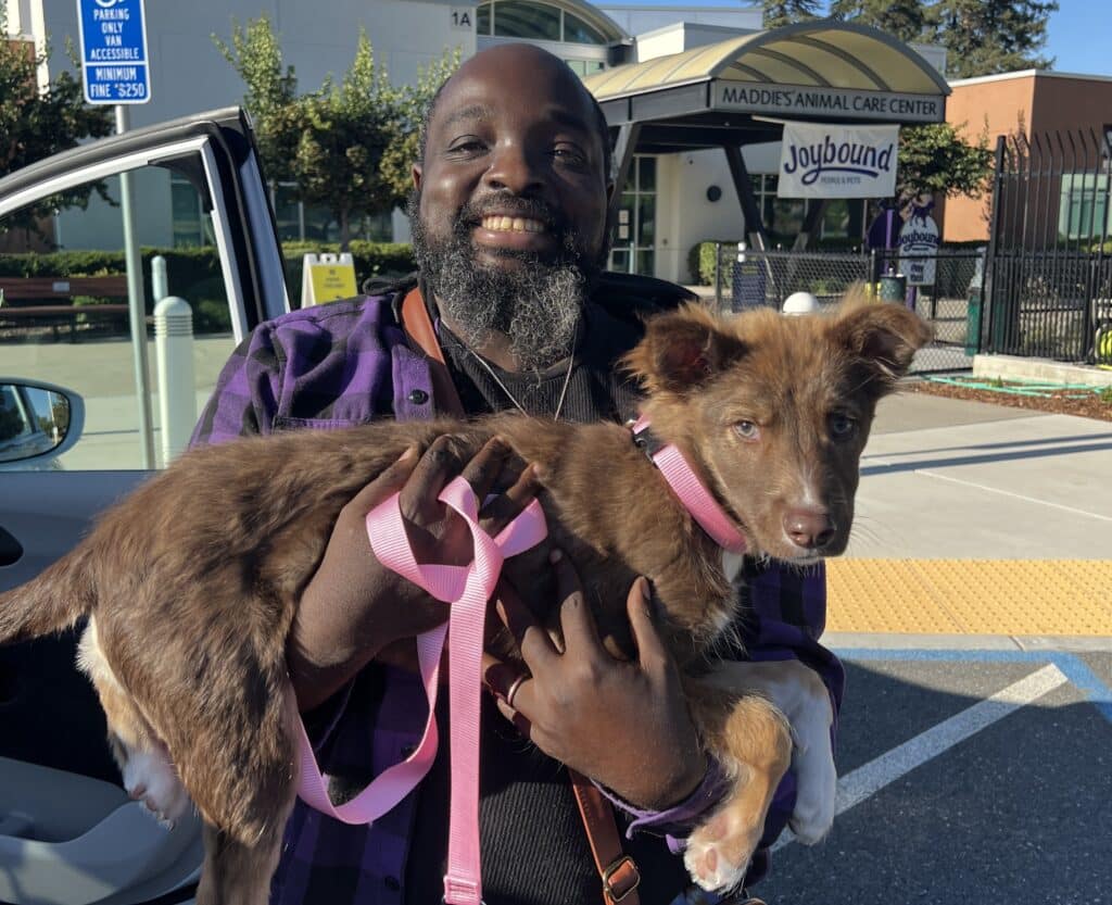 A man with a beard is smiling while holding a brown puppy with a pink leash outside an animal care center. The background features the center's entrance and signage, indicating a joyful adoption moment.