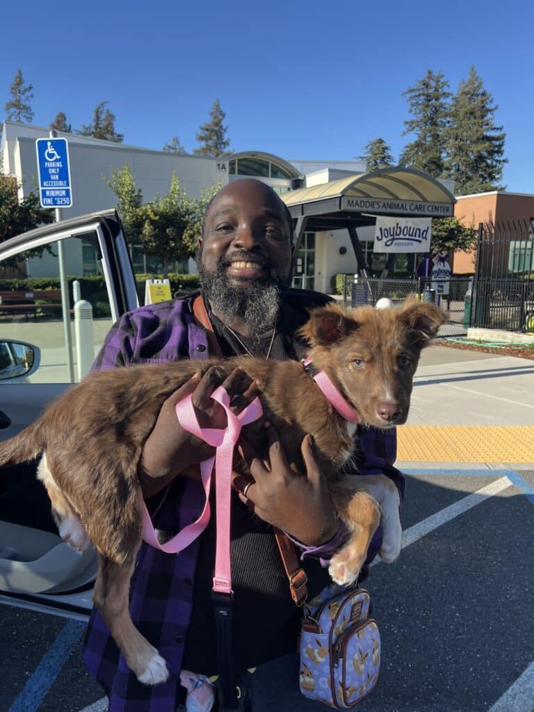 A smiling man holding a small brown and white puppy, with a pink leash, outside an animal care center. The background shows the center's sign, trees, and part of a vehicle. The scene is bright and sunny.