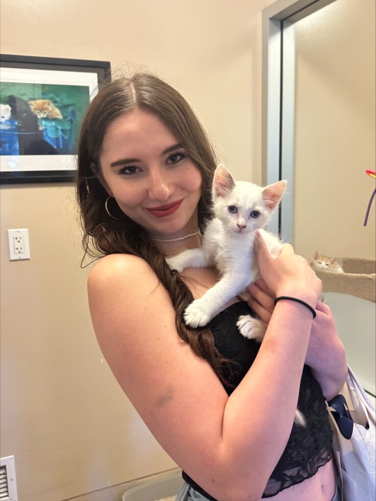 A young woman with long hair poses smiling while holding a small white kitten in her arms. The background includes a glimpse of framed artwork and room decor, representing a setting related to animal adoptions at Joybound.