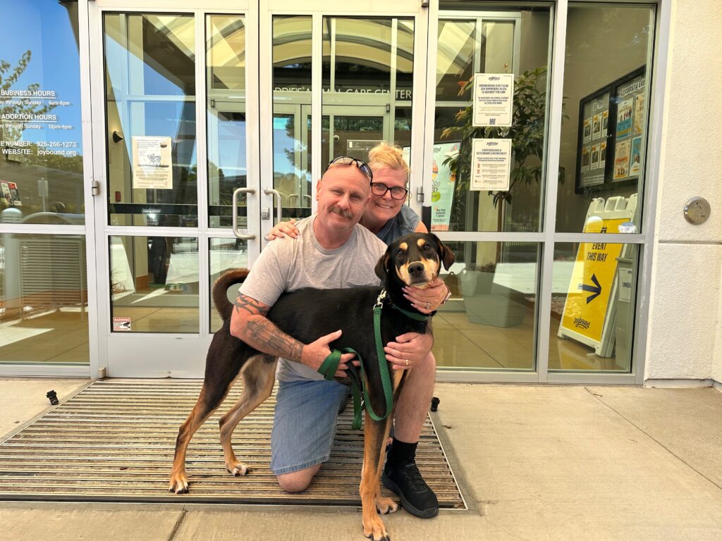 A man and a woman sit on a metal grate outside an animal shelter, smiling as they pose with a dog. The dog, on a leash, is in the man's lap. Behind them, large glass doors are visible with informational posters.