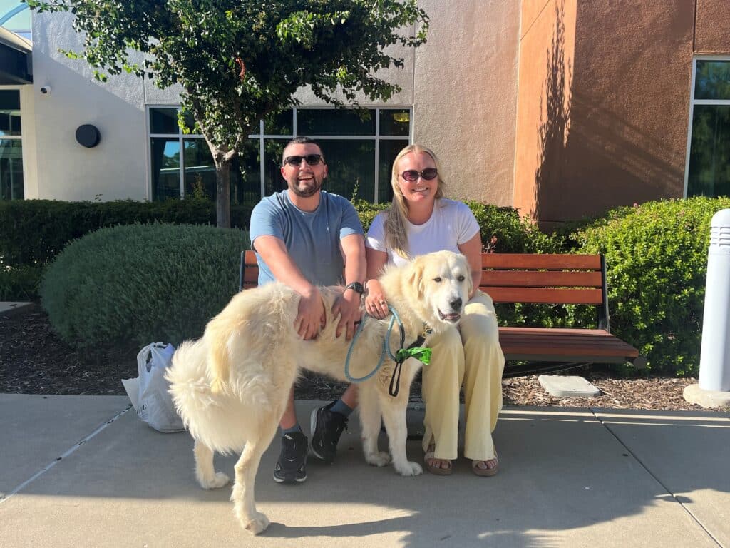 A man and a woman smile while sitting on a bench outdoors next to a large, white dog on a leash. The setting features greenery and a building in the background, suggesting a relaxed atmosphere.