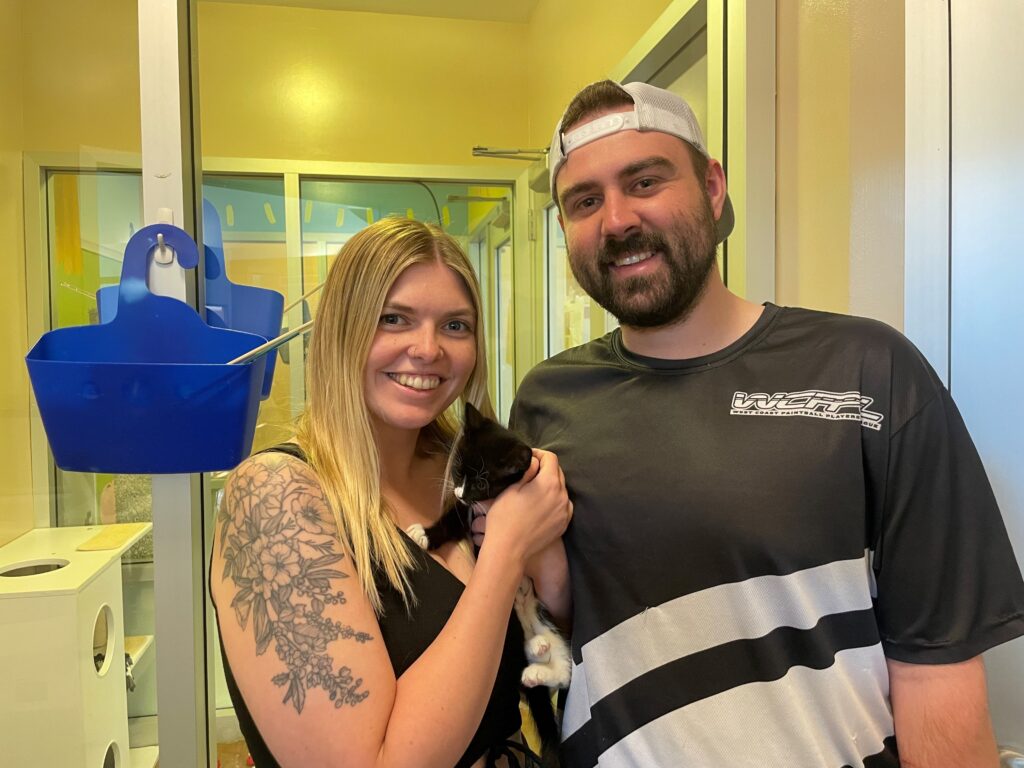 A couple stands together in a brightly lit room, smiling at the camera while holding a small black and white kitten. The woman has long blonde hair and a tattoo on her arm, while the man wears a black and white shirt.
