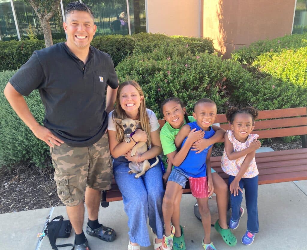 A smiling man and woman sit on a bench surrounded by four children, all posing with a small dog. The man wears a black shirt and camouflage shorts, while the woman holds the dog. The children range in ages and are wearing casual shirts.