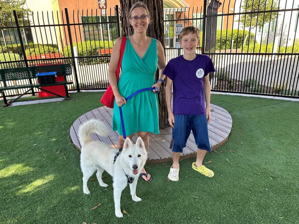 A woman in a green dress and a boy in a purple shirt stand together beside a white dog on a grassy area. The woman holds the dog's leash, and they are positioned near a wooden platform with a tree in the background.