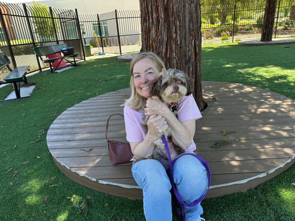A woman sitting on a wooden circular platform in a park, holding a small dog. She has long light-colored hair and is wearing a pink shirt and jeans. The background features a grassy area and a fenced space with benches.