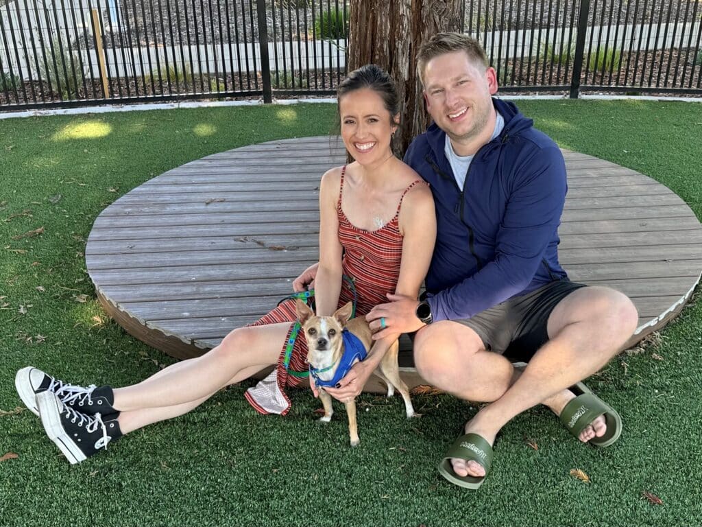 A woman in a striped dress and a man in a blue jacket sit on a circular wooden platform in a grassy area, holding a small dog wearing a blue harness. The image represents a recent pet adoption, supporting "Adoptions at Joybound."