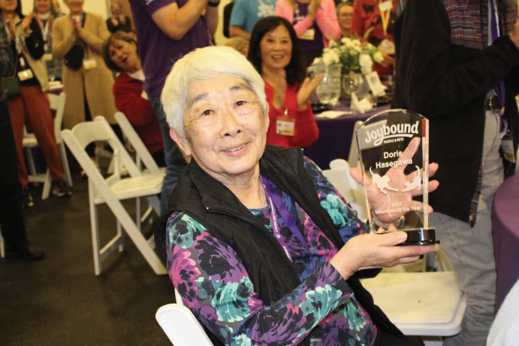 An elderly woman, Dorita Hasegawa, smiles while holding a Volunteer of the Year award at a celebratory event. In the background, attendees are clapping and celebrating in a decorated room with floral arrangements.