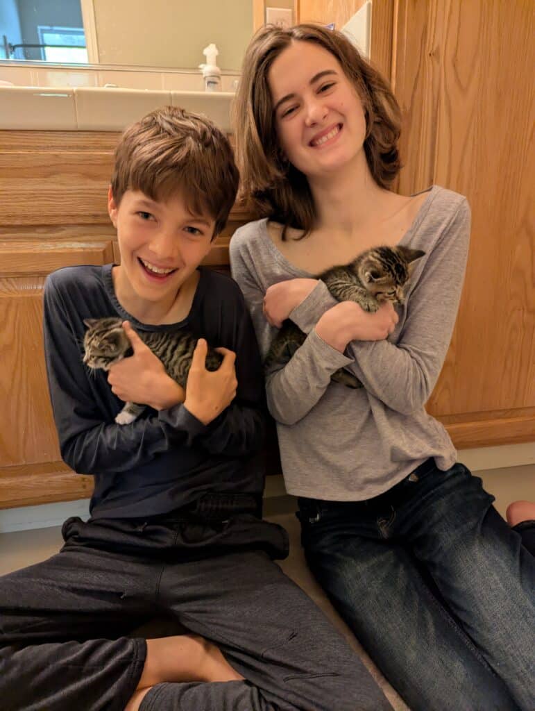 Two children sitting on a tiled floor in a bathroom setting. A boy on the left holds a small tabby kitten while smiling. A girl on the right holds a similar kitten and shows a joyful expression. Wooden cabinetry and a mirror are visible in the background.