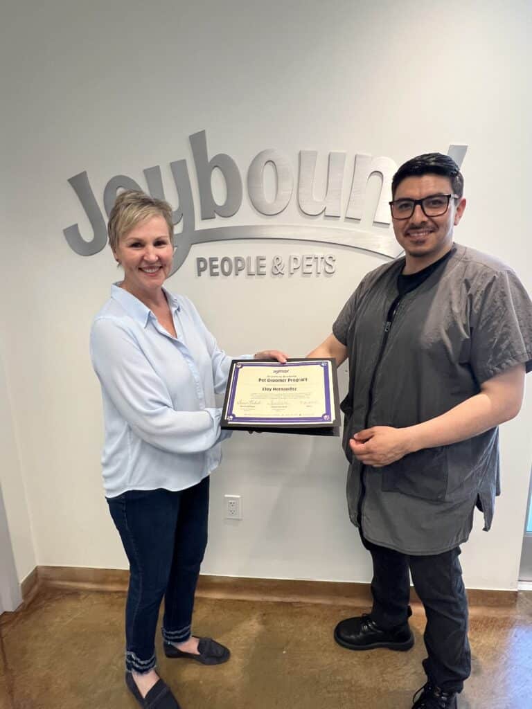A woman and a man are standing in front of a wall with the sign "Joybound People & Pets." The woman, wearing a light blue blouse, hands a certificate to the man, who is dressed in a gray shirt and glasses, smiling as they pose together.