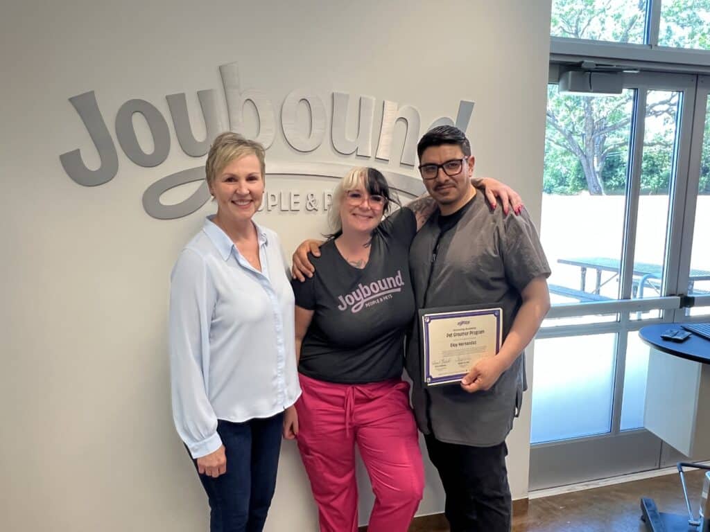 Three individuals stand together in front of a wall displaying the "Joybound" logo. The first person, wearing a blue blouse, is on the left. In the middle, a woman in a black shirt with the Joybound logo holds a certificate. The third person, on the right, wears glasses and has short hair.