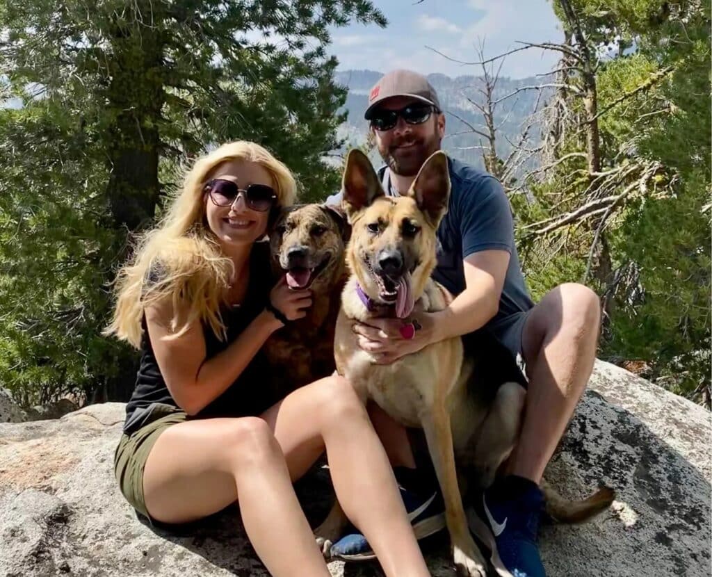 A woman with long blonde hair and sunglasses sits on a rock, holding a small brown dog. Next to her, a man in a gray shirt and cap sits with a tan and white dog. They both smile at the camera with trees and a mountainous background.
