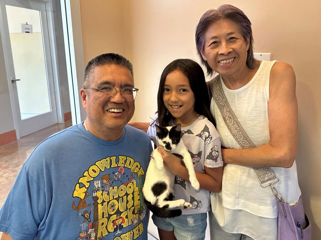 A smiling family poses for a photo in a bright room. A man and woman stand on either side of a young girl holding a black and white kitten. The setting suggests a recent adoption event, relating to "Adoptions at Joybound."