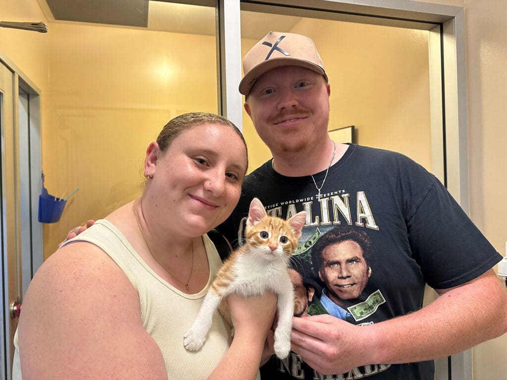 A woman and a man are holding an orange and white kitten in a pet adoption facility. The woman has light brown hair and is wearing a light tank top, while the man is wearing a black t-shirt and a beige cap. This image relates to "Adoptions at Joybound."