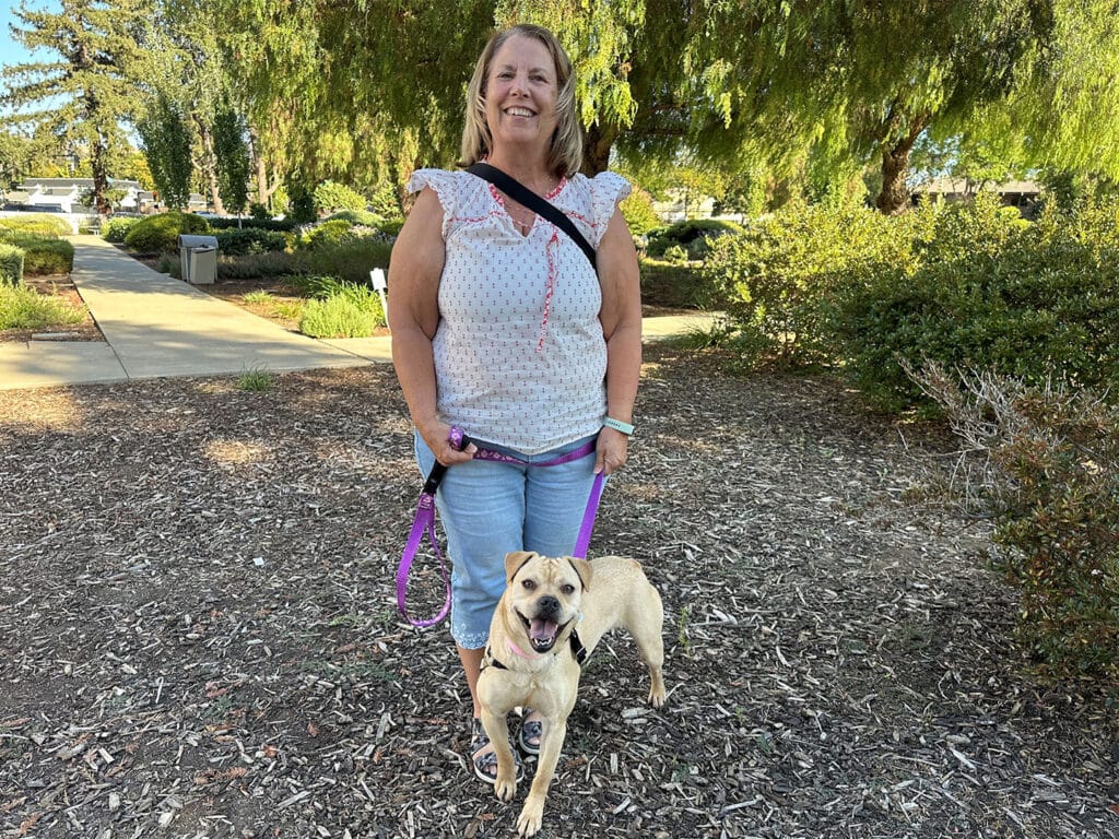 A woman stands on a path in a park, smiling and holding a leash attached to a light-colored dog. The park features greenery and well-maintained landscaping. This image relates to adoptions at Joybound.