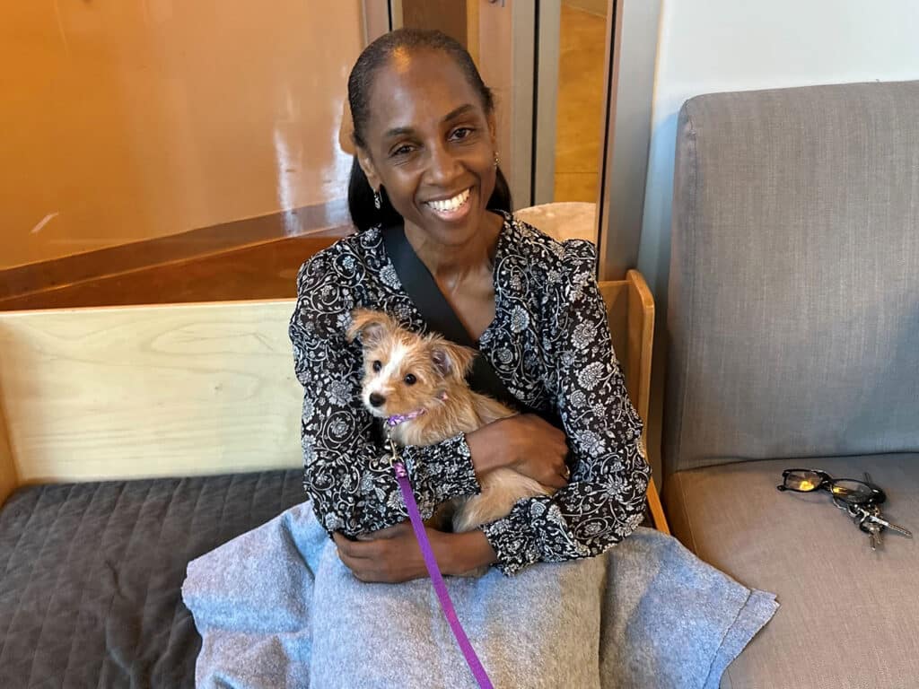 A woman holding a small dog on her lap, seated on a bench in a casual indoor setting. She is smiling and wearing a black and white floral blouse. The scene represents adoptions at Joybound, highlighting the bond between pets and their new owners.