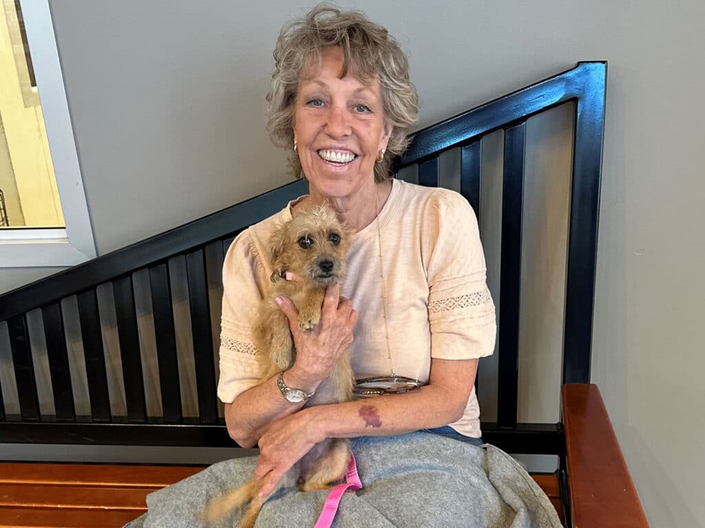A woman sitting on a bench holding a small brown dog, smiling. The setting appears to be an indoor area with a wooden bench and a decorative wall behind her. This image is part of the 'Adoptions at Joybound' recap for the week of August 4.