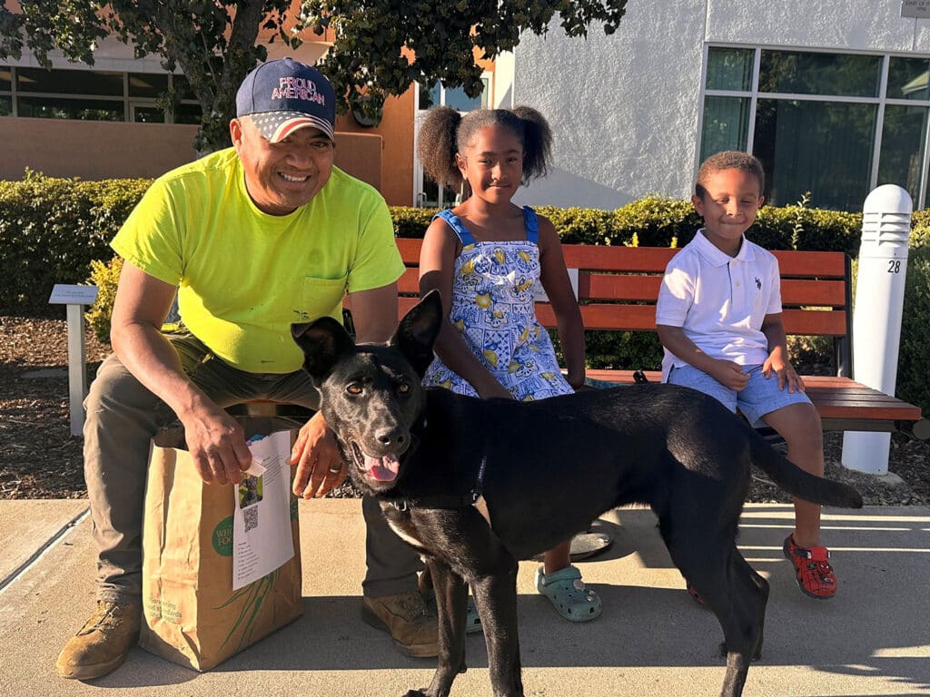 A man in a bright yellow shirt sits with a black dog at his feet, next to two children on a bench. One child wears a blue floral dress and the other a white shirt, both are smiling. The photo represents Adoptions at Joybound.