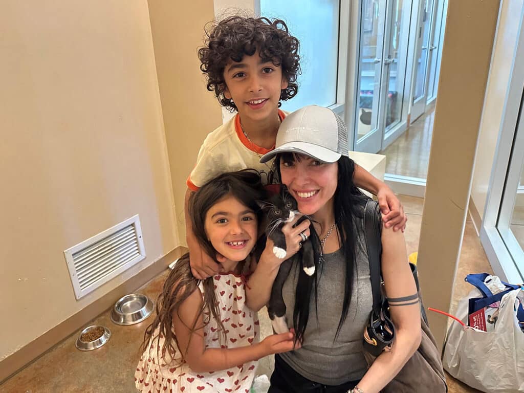 A woman with long dark hair holds a kitten while posing with two children, a boy and a girl, in a brightly lit room. The boy, with curly hair, stands behind her, and the girl, dressed in a dress with hearts, is beside her. This image relates to Adoptions at Joybound.