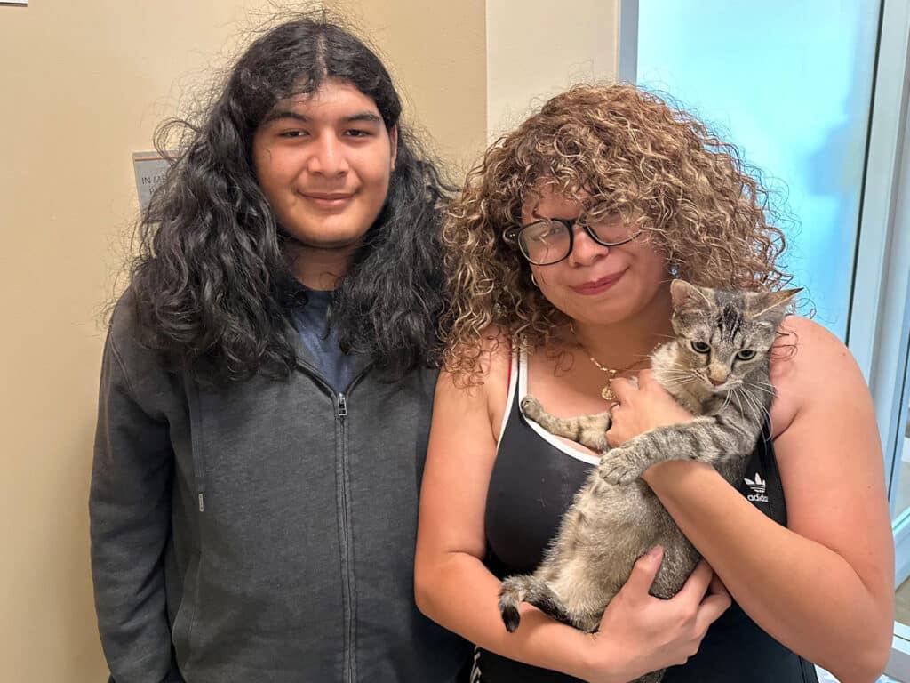 A young man with long dark hair and a gray hoodie stands next to a young woman with curly hair holding a gray tabby cat. They are posing in a well-lit room, smiling slightly, after completing their adoption process at Joybound.