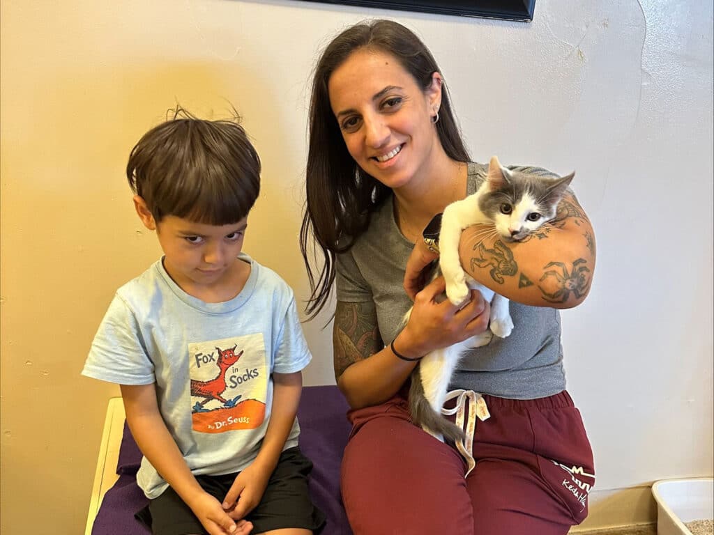 A woman holding a gray and white kitten on her arm sits beside a young boy looking slightly disgruntled. They are in a cozy indoor space, likely a shelter. The image highlights the joy of pet adoption, emphasizing Adoptions at Joybound.