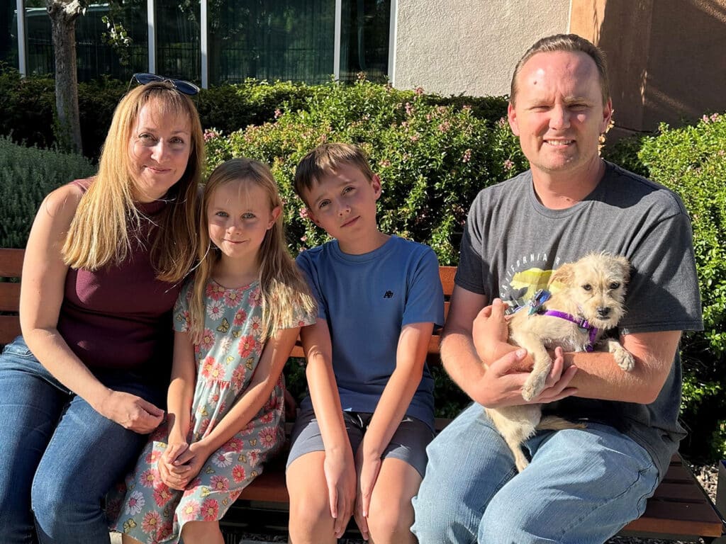 A family of four sitting on a bench outside, with a woman, a girl in a floral dress, a boy wearing a blue shirt, and a man holding a small dog. The background features greenery and a building, emphasizing the theme of adoptions at Joybound.