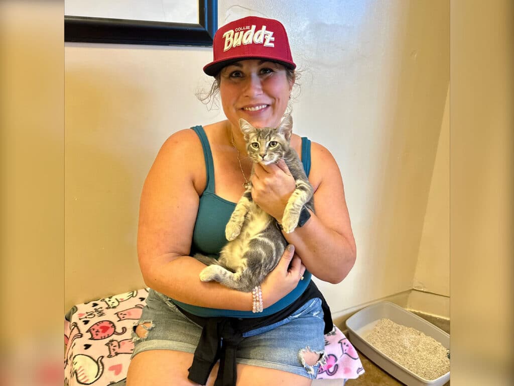A woman wearing a red cap with "Buddz" sits on a blanket with a playful gray tabby kitten in her arms. The background features a litter box and neutral walls, suggesting an adoption environment. This scene illustrates "Adoptions at Joybound".