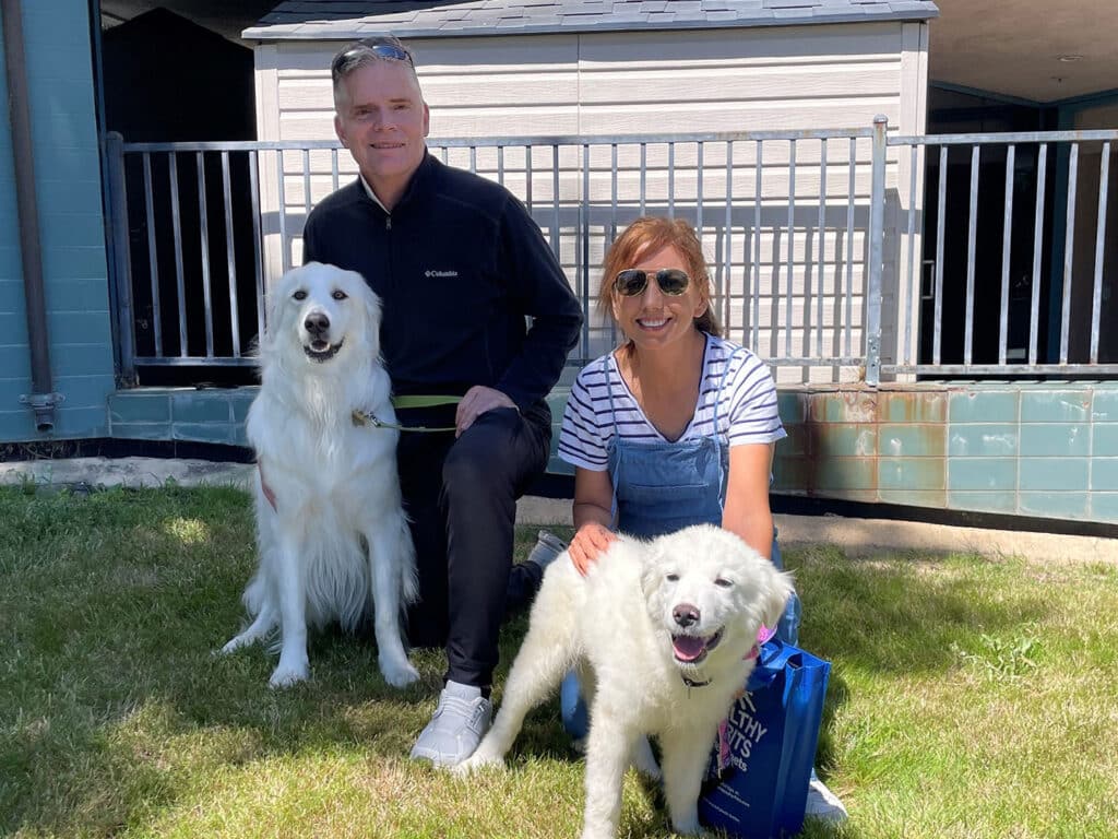 A man and a woman are sitting on the grass outside a building, each with a fluffy white dog. The woman wears sunglasses and a striped shirt, while the man is dressed in a black jacket. This image highlights Adoptions at Joybound.