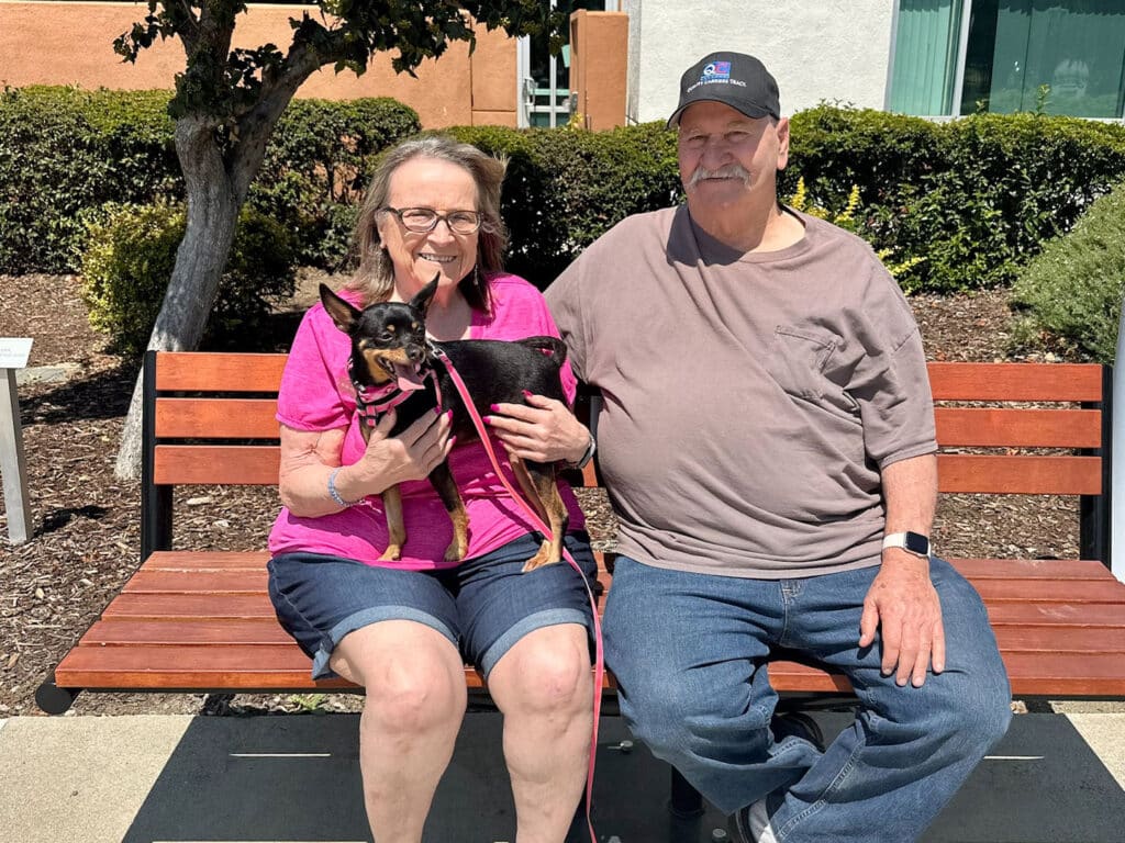 An older woman and man sitting on a wooden bench outdoors, smiling while holding a small black and tan dog on a leash. The background features greenery and a building, indicating a welcoming environment. Relevant to Adoptions at Joybound.
