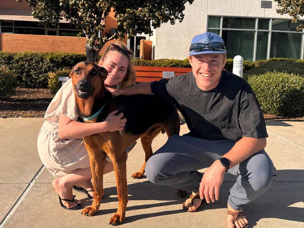 A woman and a man pose beside a brown dog in a sunny outdoor setting. The woman, wearing a light dress, gently holds the dog, while the man smiles beside them. The background features a building and landscaped areas, highlighting the adoption theme connected to 'Adoptions at Joybound'.