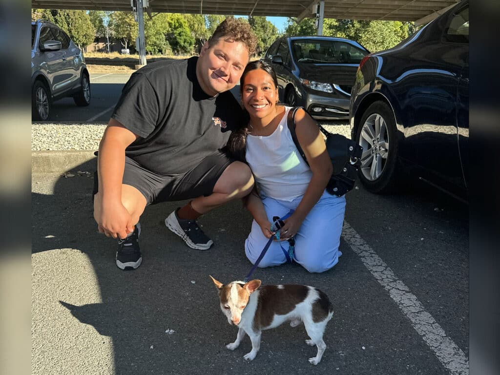 A man and a woman smile while kneeling beside a small dog in a parking area. The man is wearing a black t-shirt and shorts, while the woman is in a white top and light blue pants. The image captures a moment of joy, highlighting adoptions at Joybound.