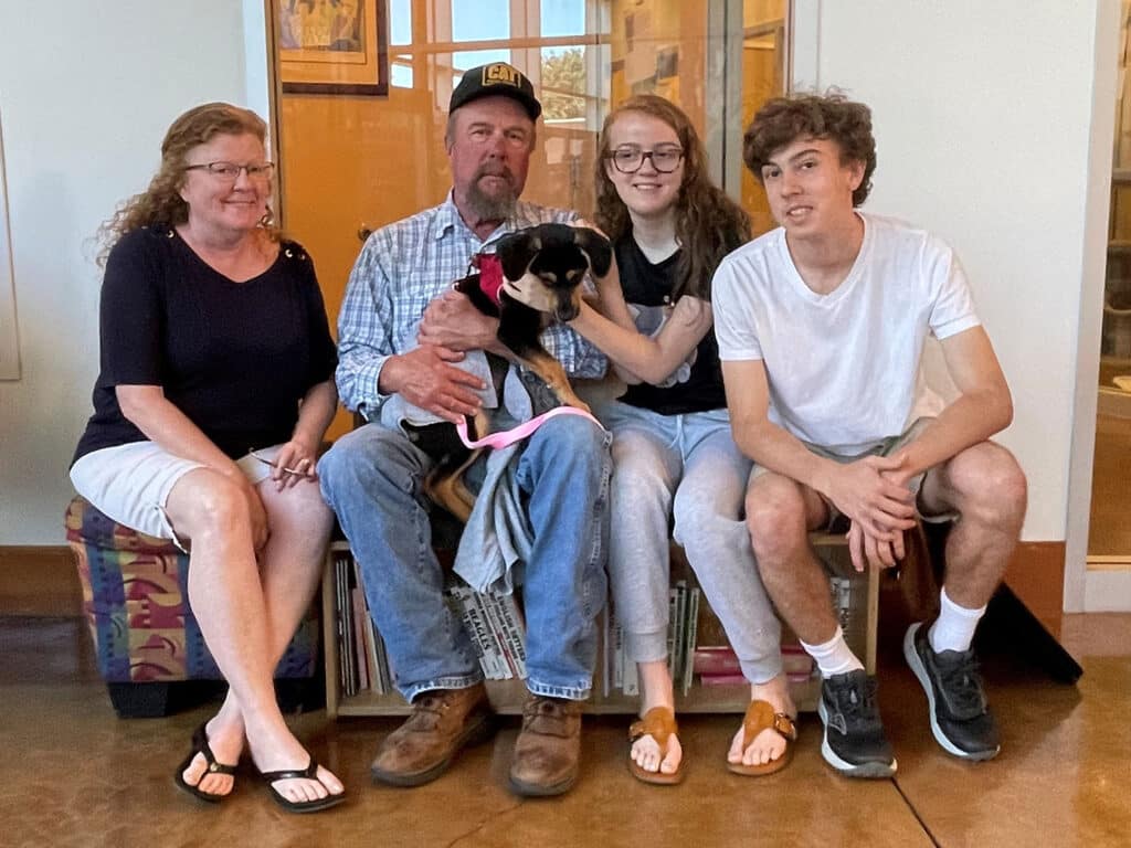 A family of four sits together on a bench, with a young woman holding a small dog in her lap. A man in a hat smiles next to her, while two young adults sit at either end. The setting appears to be an adoption center, highlighting "Adoptions at Joybound".