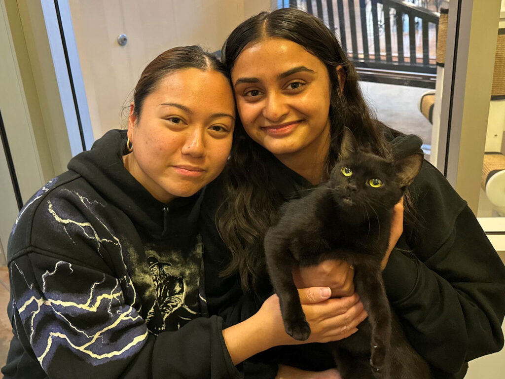 Two young women, smiling while holding a black cat between them, in a bright room with wooden furniture. This image highlights successful adoptions at Joybound, capturing a moment of joy and companionship.