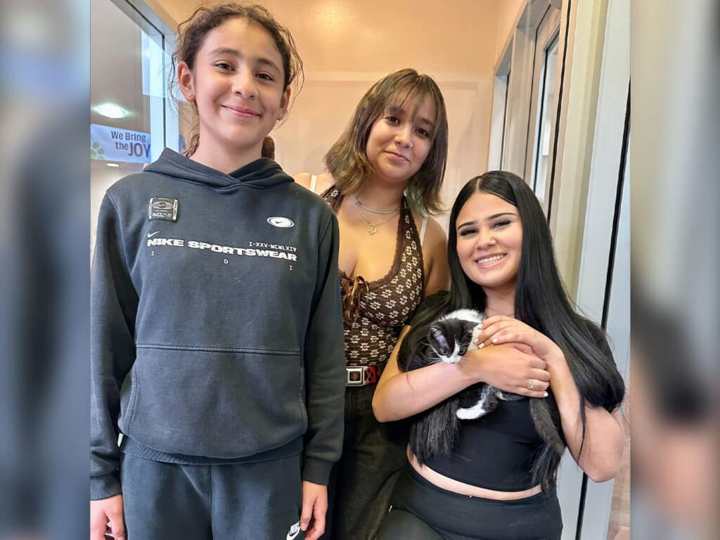 Three young individuals smiling together in a brightly lit corridor. One girl holds a small black and white kitten, while the others stand beside her. This image is related to "Adoptions at Joybound" and showcases a positive adoption moment.