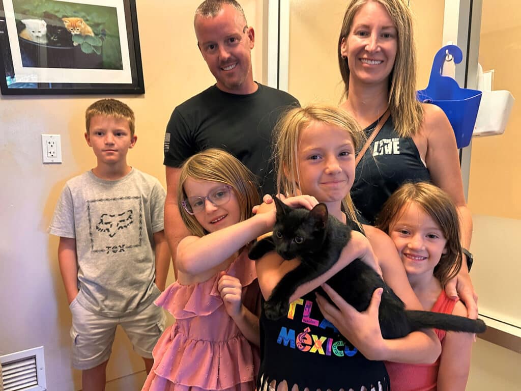 A family of five standing together inside an animal shelter. Two girls are holding a black kitten, while the parents and two other children smile. This image is related to the theme of "Adoptions at Joybound" following their recent adoption week.