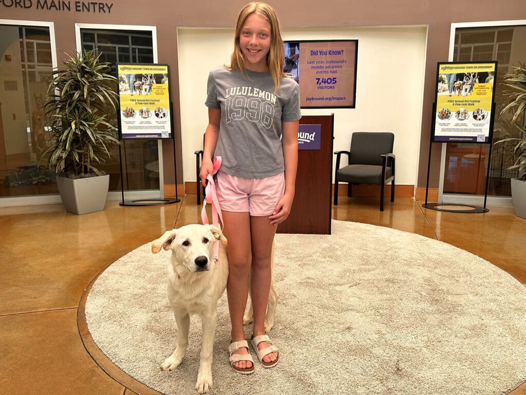 A young girl stands with a cream-colored dog on a circular rug in an adoption center. The girl wears a gray t-shirt and pink shorts, holding a leash. Supportive banners are visible in the background, highlighting "Adoptions at Joybound".