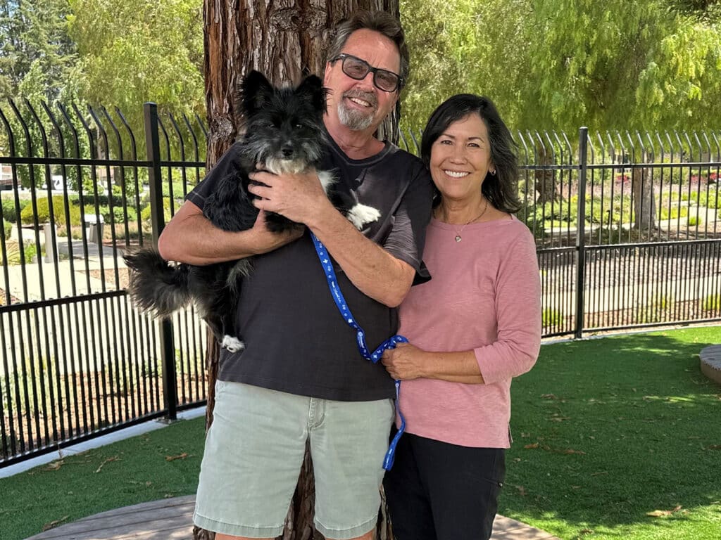 A man holding a small black dog and a woman standing beside him, both smiling. They are outdoors in a park-like setting with green grass and trees in the background. This image relates to Adoptions at Joybound.