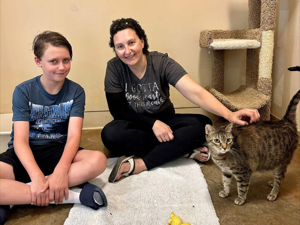 A boy and a woman sitting together on the floor of an animal shelter. The boy wears a blue shirt and black shorts, while the woman has short hair and is touching a small brown tabby cat. A scratching post is visible in the background. Adoptions at Joybound.