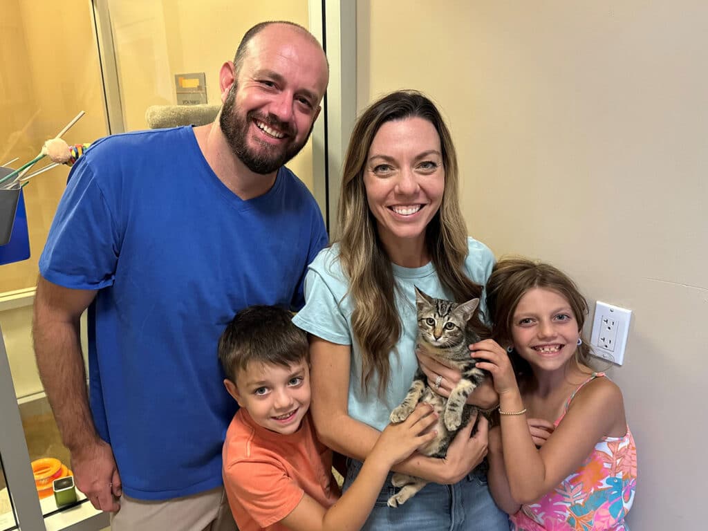 A family of four poses together in a bright room. The father, wearing a blue shirt, stands on the left, while the mother, in a light blue top, holds a small tabby cat. Two children, a boy and a girl, stand beside them, smiling as they point at the cat. This image highlights recent adoptions at Joybound.