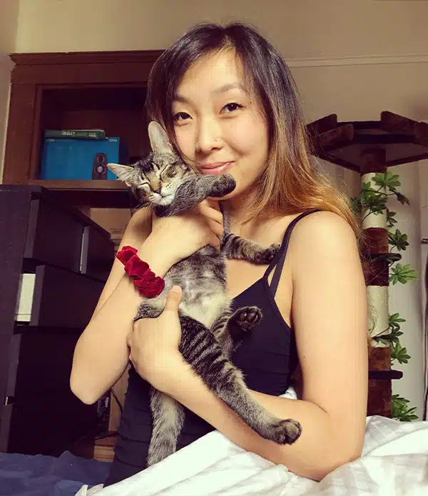A woman in a black tank top holding a small tabby cat in her arms, sitting on a bed. The background shows a bookshelf and a cat tower, with greenery visible in the room. The woman has shoulder-length hair and a relaxed expression.