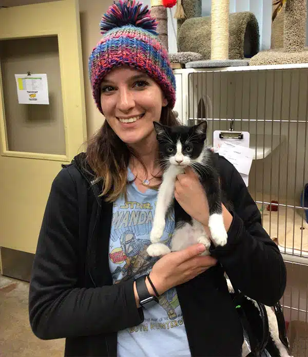 A woman wearing a colorful knit hat holds a small black and white kitten in a pet adoption center. The background shows shelves with cat supplies and another cat tower.
