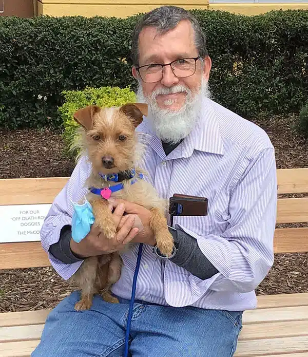 An elderly man with glasses and a white beard sits on a bench, holding a small brown dog with a blue ribbon around its neck. Behind them, there is greenery and a sign partially visible in the background.