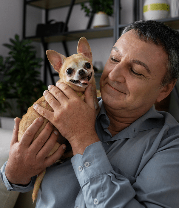 A man in a blue shirt holding a small tan Chihuahua in his arms, both looking content. The setting includes green plants and stylish shelving in the background. This image relates to the Joybound donate items campaign.