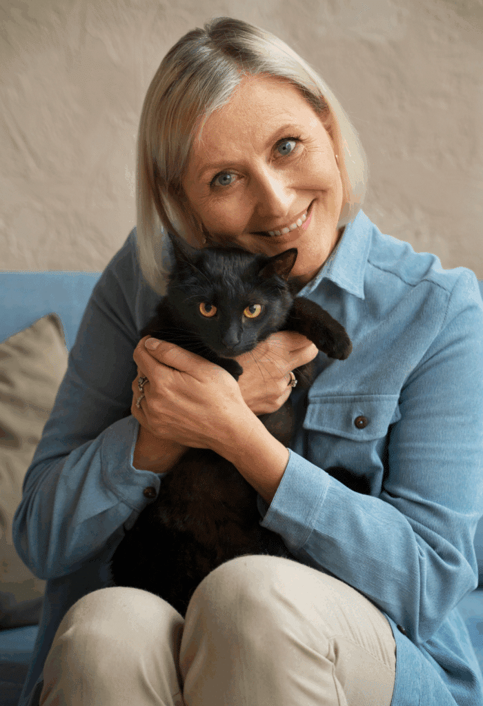 A smiling elderly woman holding a black cat close to her chest, sitting on a blue couch in a cozy room. The woman's hair is silver, and she wears a blue button-up shirt. This image represents the joy of companionship, related to Joybound donate items.